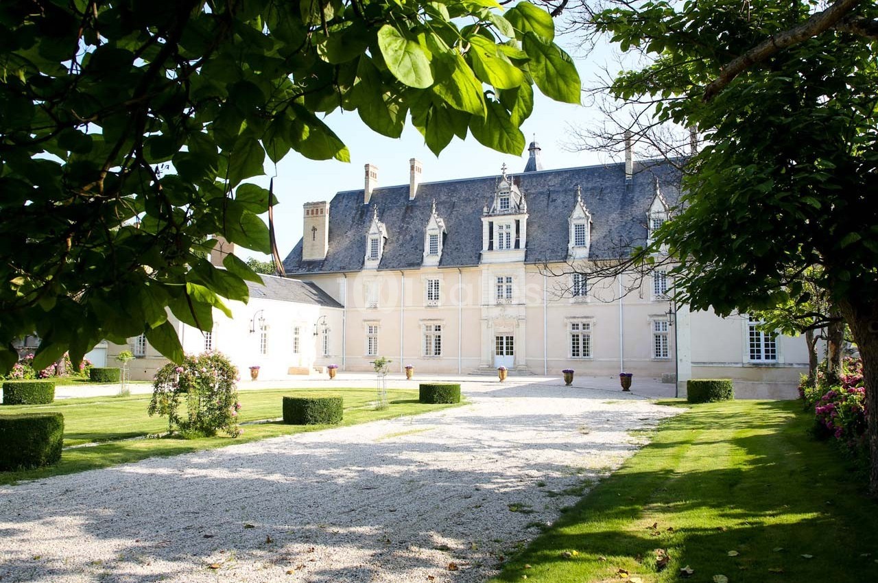 Façade d'un manoir historique entouré d'un jardin verdoyant avec allée gravillonnée et arbustes taillés.