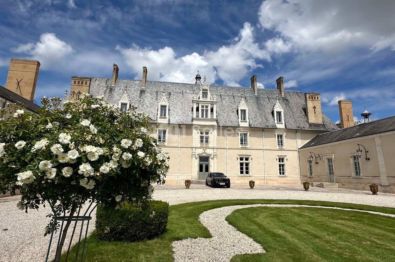 Façade d'un manoir en pierre avec toit en ardoise, jardin aménagé au premier plan et ciel partiellement nuageux.