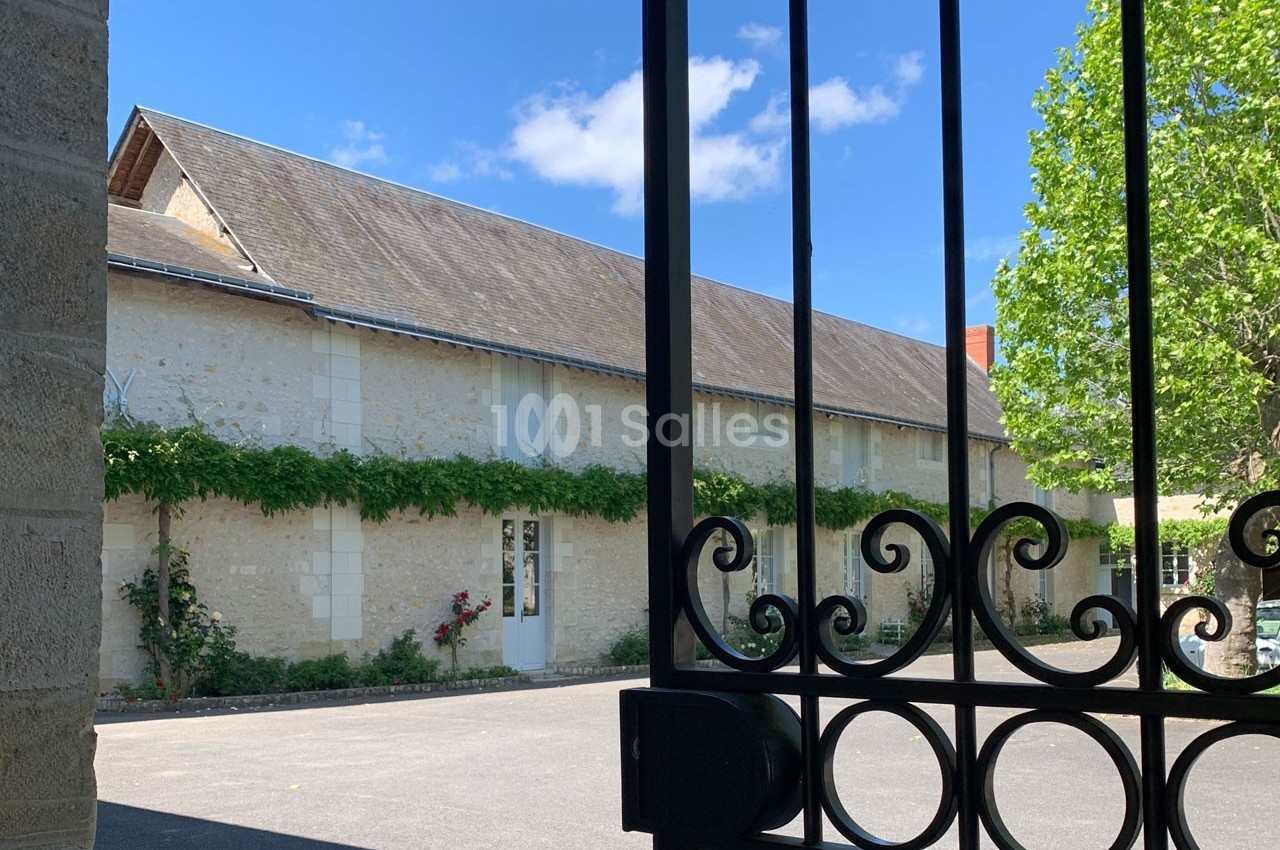 Bâtiment en pierre clair avec toit en pente, entouré de verdure, vu à travers une grille en fer forgé.