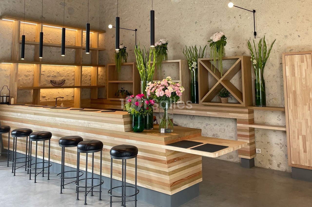 Comptoir en bois avec tabourets noirs, étagères décorées de fleurs et éclairage suspendu dans un espace moderne.
