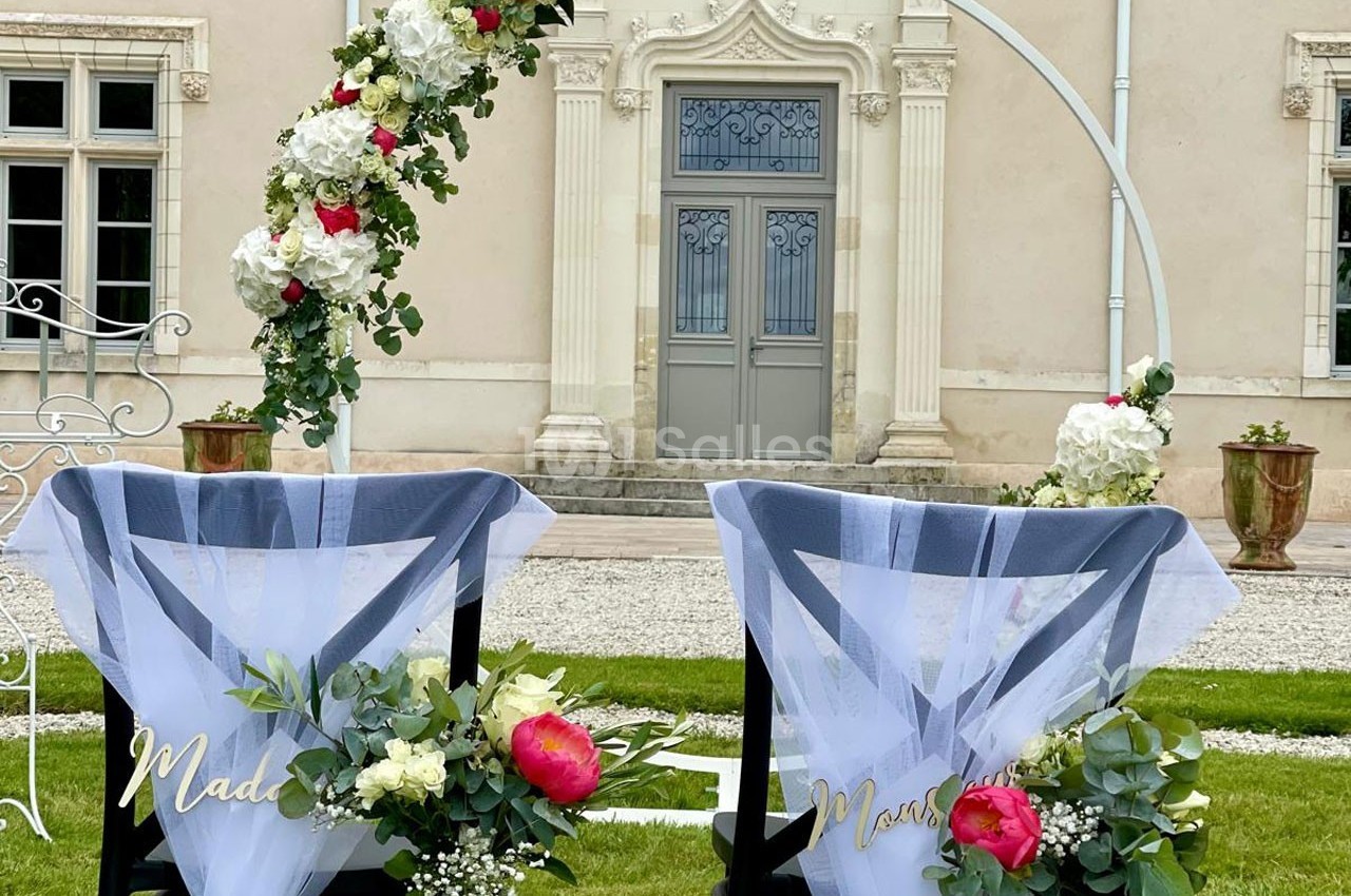 Deux chaises décorées de voiles blancs et de fleurs devant une arche florale, dans un jardin près d'un bâtiment ancien.