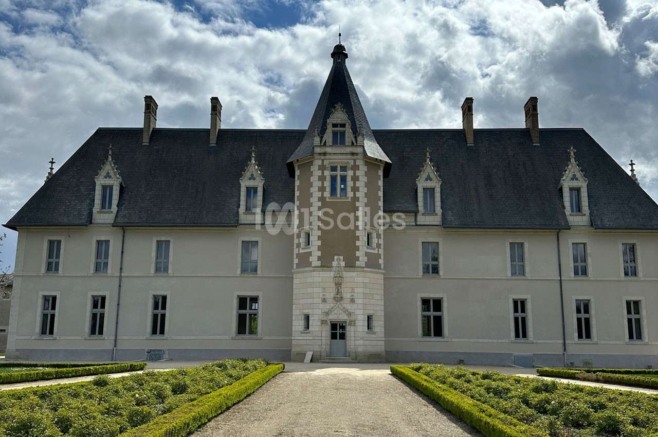 Façade d'un château avec une tour centrale, entouré de jardins symétriques sous un ciel partiellement nuageux.