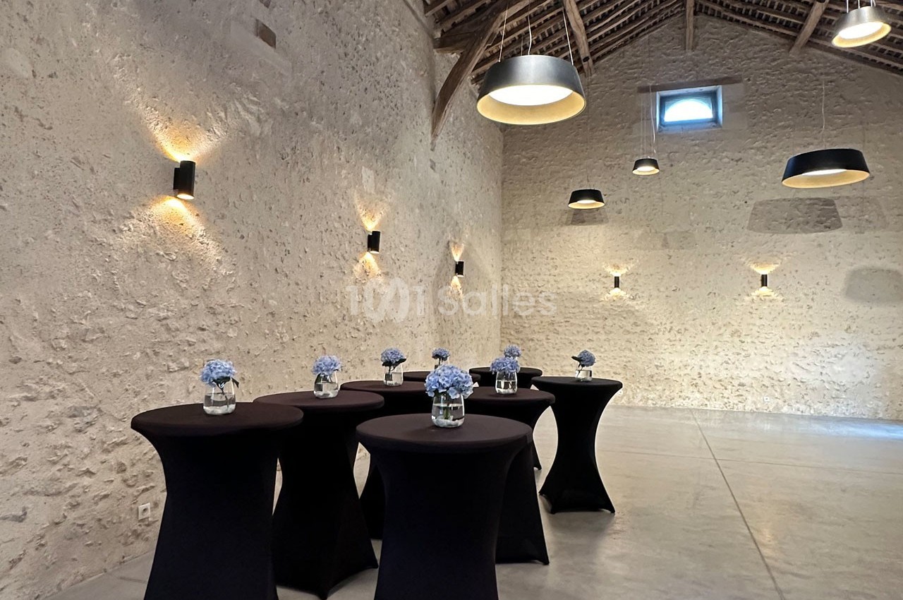 Salle en pierre avec tables hautes noires décorées de bouquets de fleurs bleues, éclairée par des suspensions modernes.