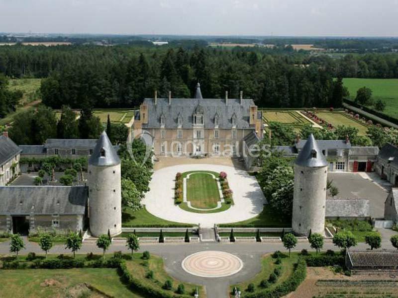 Vue aérienne d'un château entouré de jardins, de tours rondes et de bâtiments annexes, au milieu d'une campagne verdoyante.