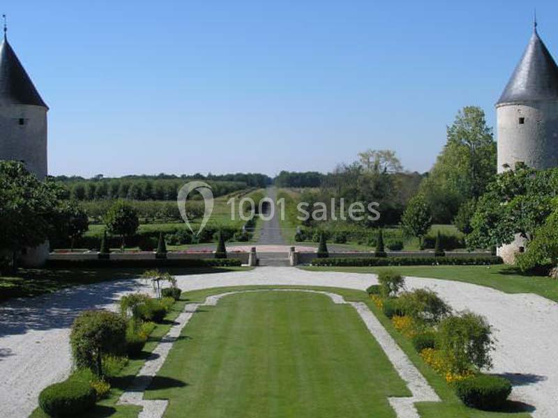Allée centrale bordée de pelouses et de parterres fleuris, encadrée par deux tours dans un paysage verdoyant.