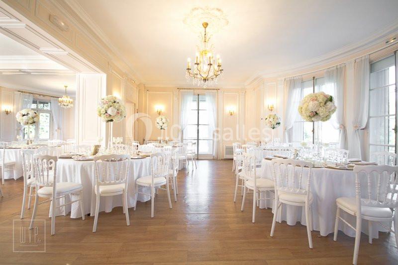 Salle de réception lumineuse avec tables rondes décorées de fleurs blanches et chaises blanches, parquet au sol.