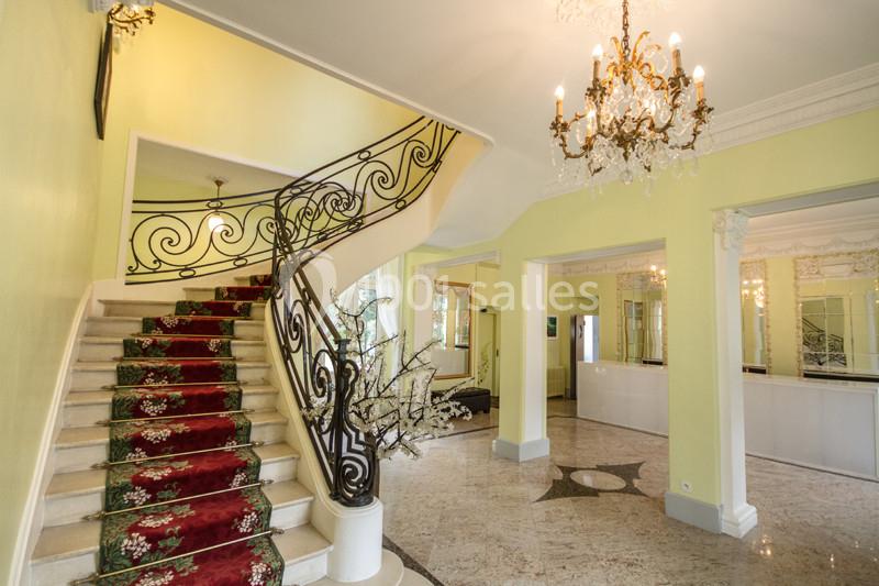Escalier avec rampe en fer forgé, tapis rouge à motifs, lustre en cristal et murs jaunes dans un hall lumineux.