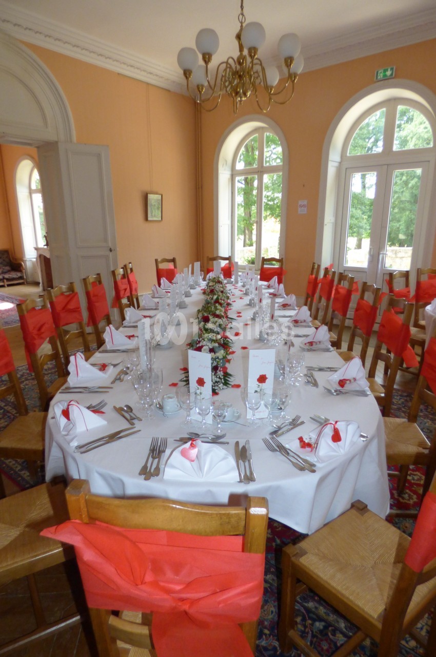 Table dressée pour un repas festif dans une salle lumineuse, avec chaises décorées de nœuds rouges et vaisselle élégante.