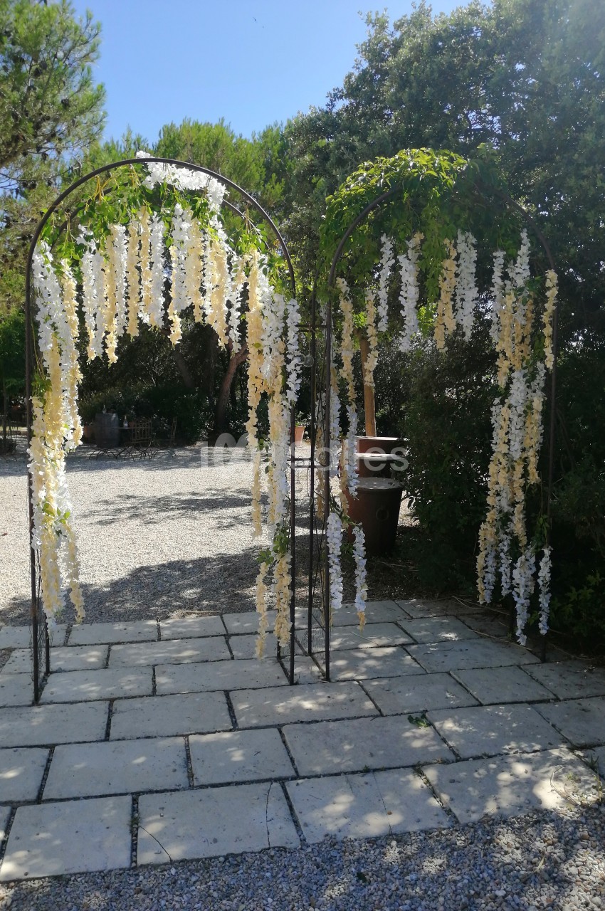 Arche métallique décorée de fleurs blanches suspendues, installée sur un sol pavé dans un jardin ensoleillé.