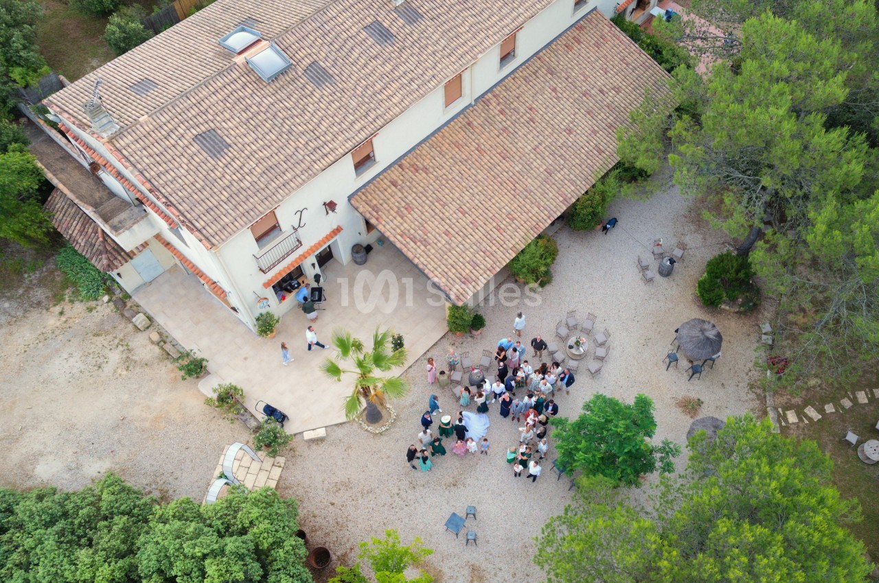 Vue aérienne d'une maison avec toit en tuiles, terrasse animée par un rassemblement de personnes et jardin arboré.