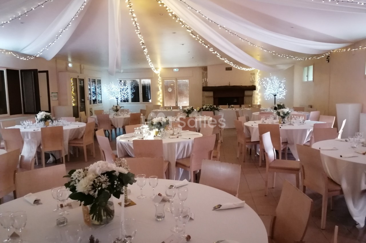Salle de réception décorée avec des tables rondes, nappes blanches, fleurs et guirlandes lumineuses suspendues.
