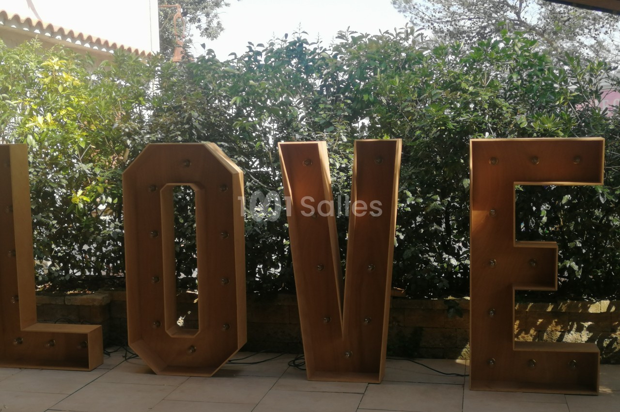 Lettres géantes en bois formant le mot ’LOVE’, éclairées par des ampoules, devant une haie verdoyante.