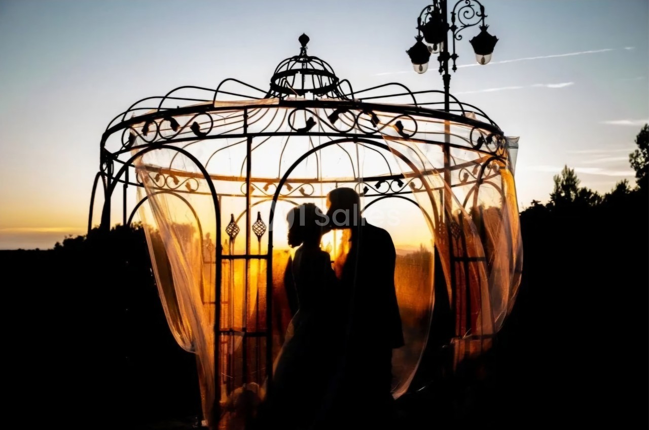 Un couple se tient sous une structure en fer forgé au coucher du soleil, entouré de voilages translucides.