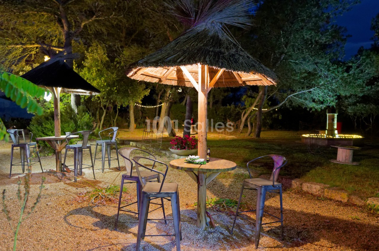 Espace extérieur éclairé avec tables en bois, chaises métalliques et parasols en paille, entouré de végétation.