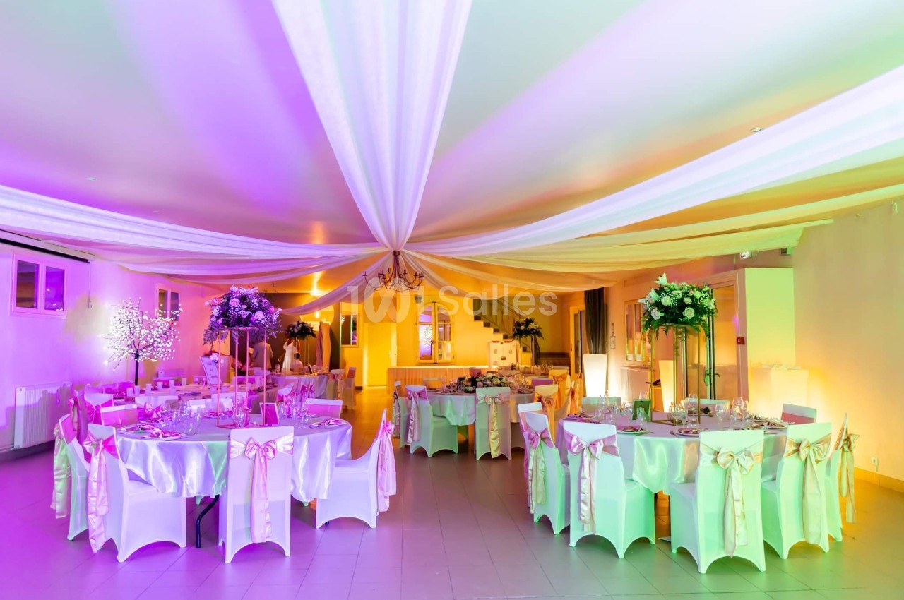 Salle de réception décorée avec des tables rondes, nappes blanches, rubans roses et éclairage coloré.