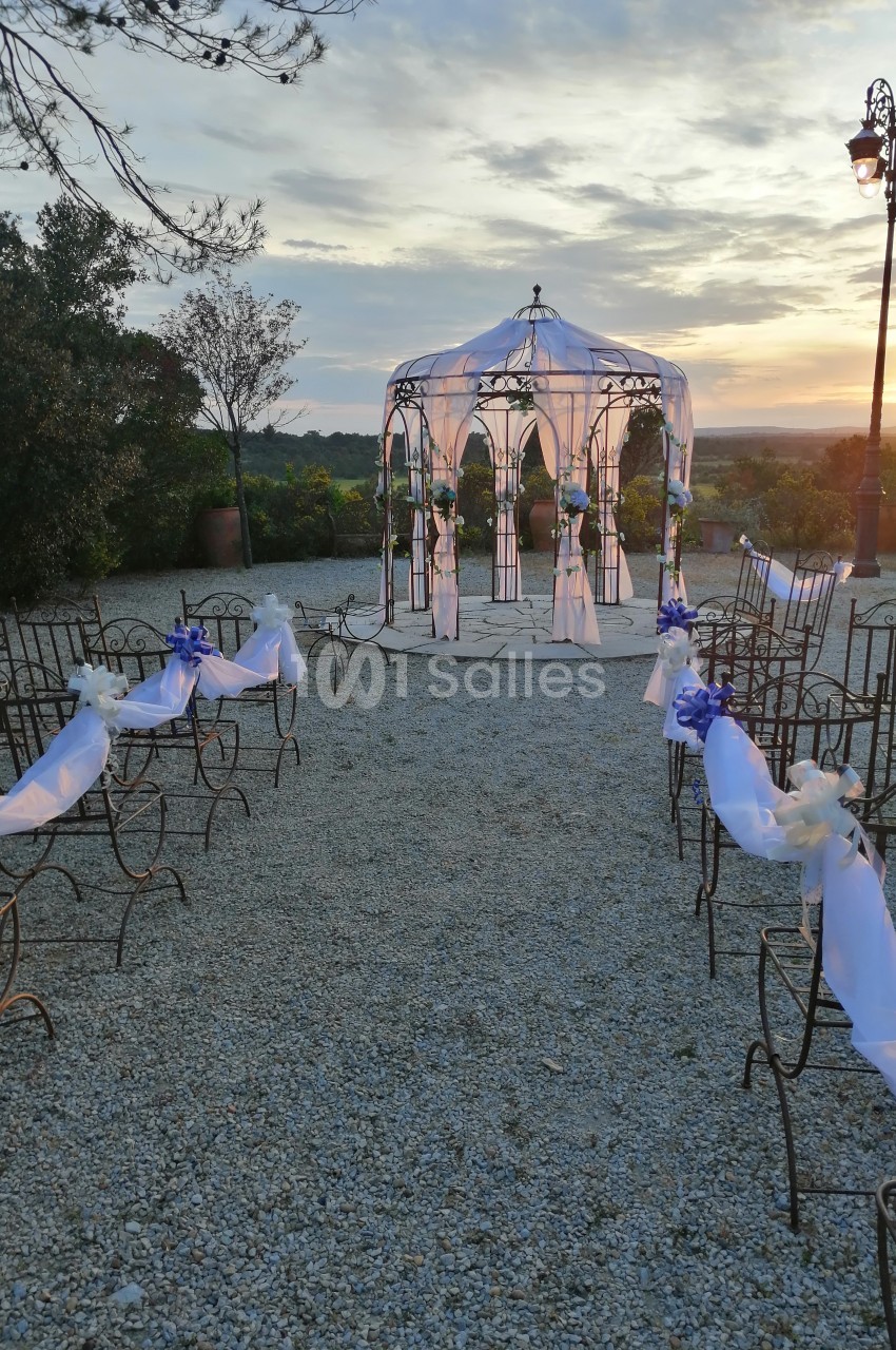 Allée décorée menant à une arche de mariage ornée de voiles blancs et de rubans bleus, au coucher du soleil.