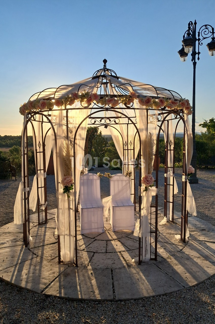 Tonnelle en fer forgé décorée de voilages et de fleurs, éclairée par le soleil couchant dans un jardin.