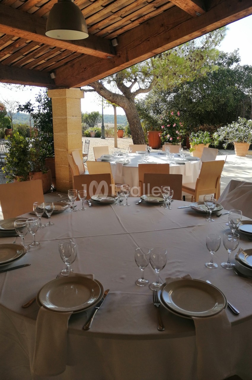 Tables rondes dressées avec nappes blanches, assiettes et verres, sous une terrasse en bois avec vue sur un jardin.