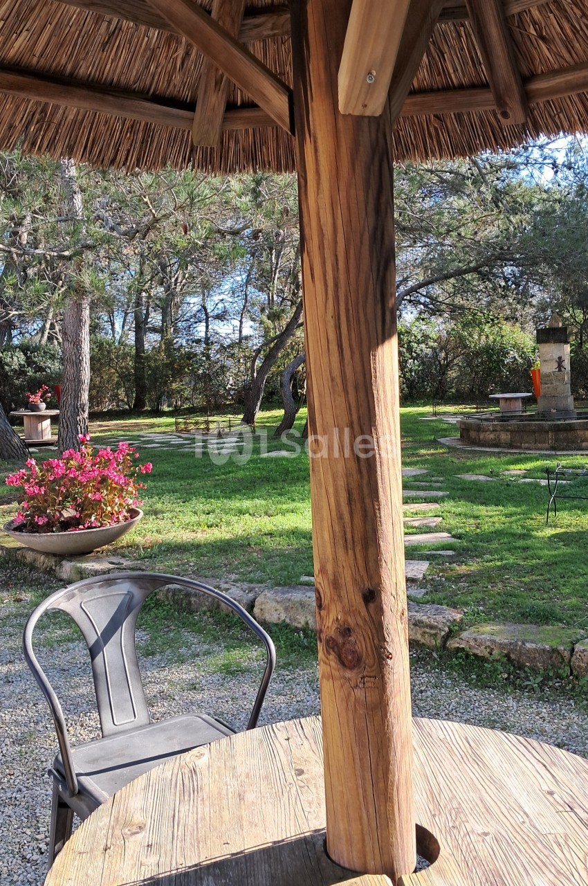 Table en bois sous un parasol en chaume, chaise métallique et jardin arboré avec fleurs et fontaine en arrière-plan.