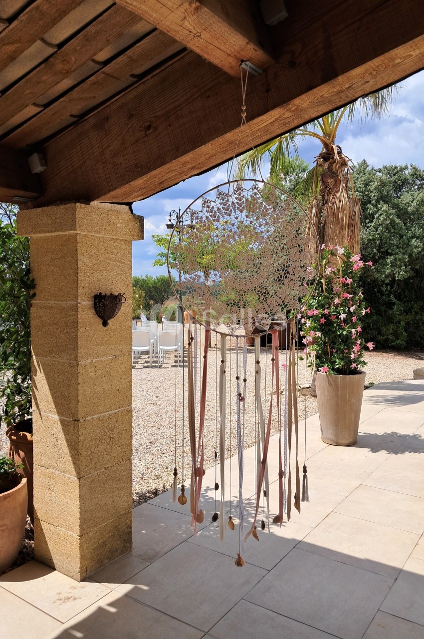 Attrait alt : Grand attrape-rêves suspendu sous une pergola en bois, entouré de plantes et d'une terrasse ensoleillée.