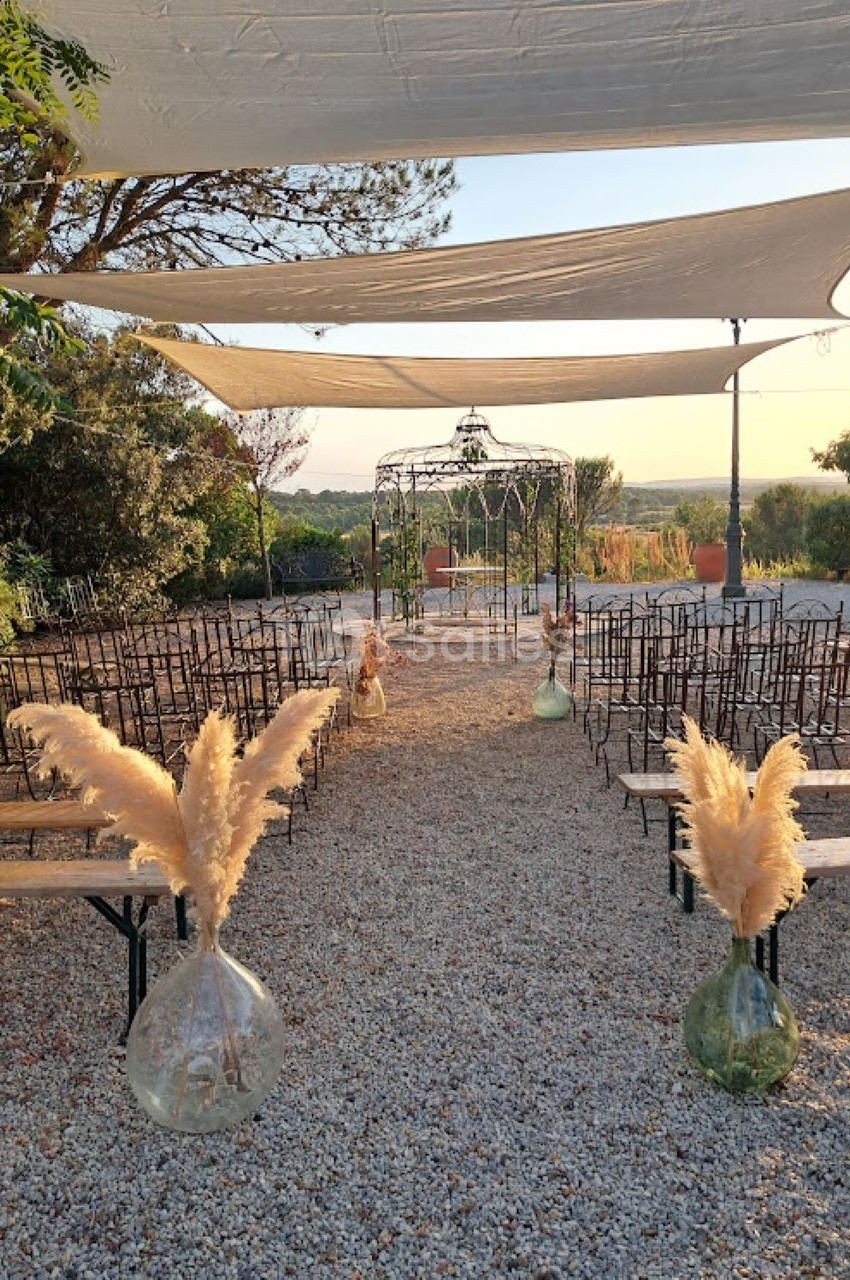 Allée centrale avec des chaises alignées, décorée de vases et de pampas, menant à une arche sous des voiles tendues.