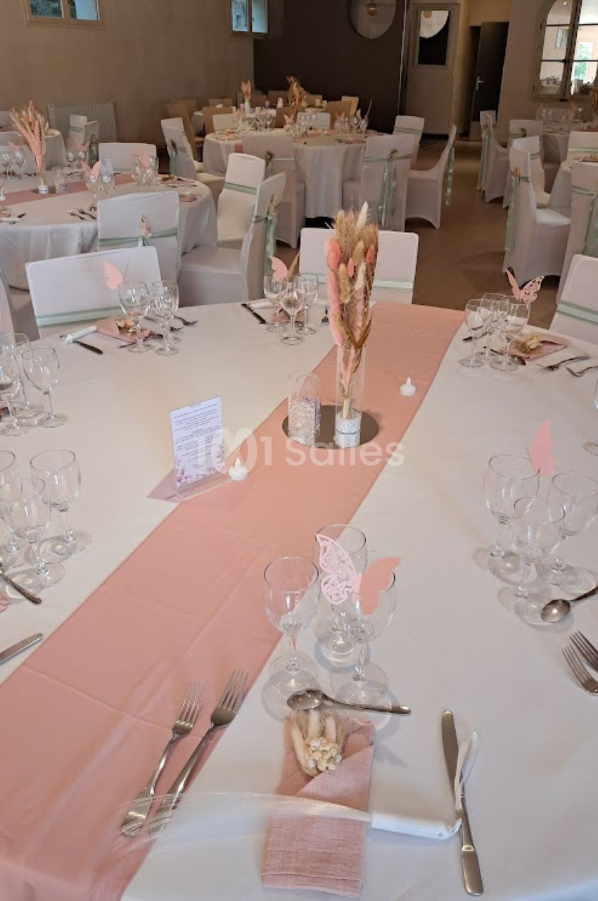 Salle de réception décorée avec des tables rondes, nappes blanches, chemins de table roses et décorations florales.