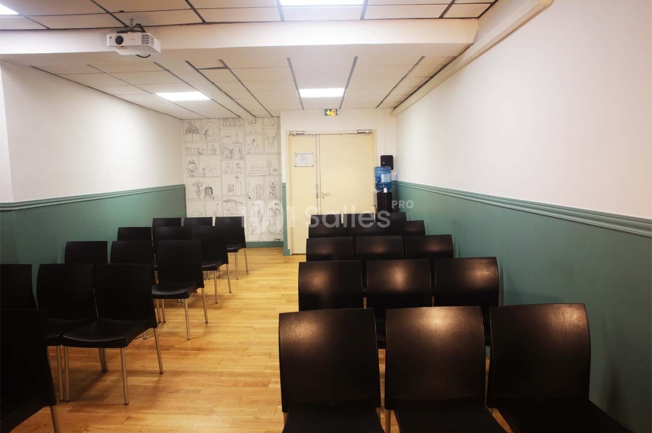 Salle de réunion avec rangées de chaises noires, sol en parquet, murs blancs et verts, et un projecteur au plafond.