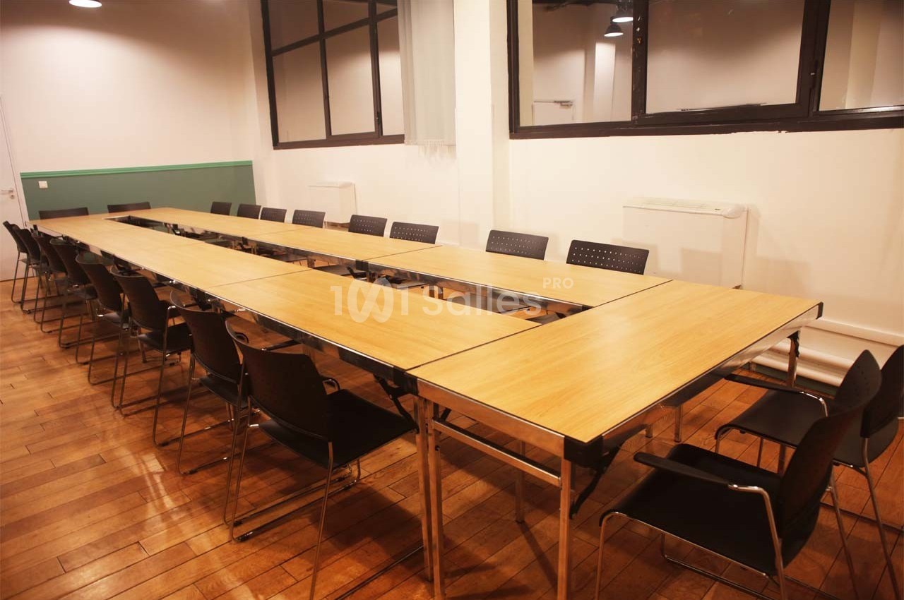Salle de réunion avec une grande table en bois en forme de U entourée de chaises noires, sol en parquet.