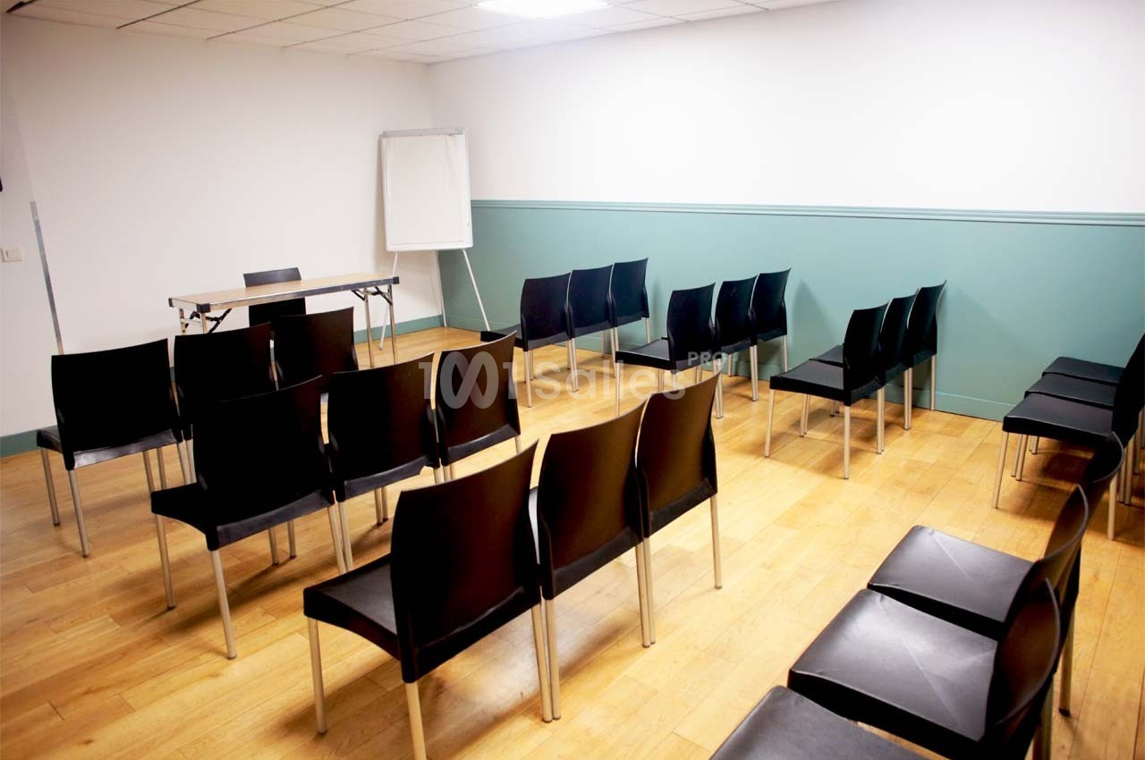 Salle de réunion avec des chaises noires alignées, un tableau blanc et une table sur un sol en parquet clair.