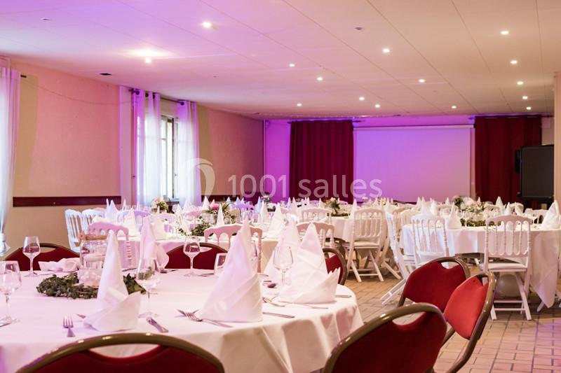 Salle de réception décorée avec des tables rondes, nappes blanches, serviettes pliées et éclairage tamisé.