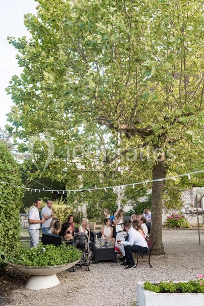 Groupe de personnes assises et debout dans un jardin ombragé, entouré de verdure et décoré de guirlandes.