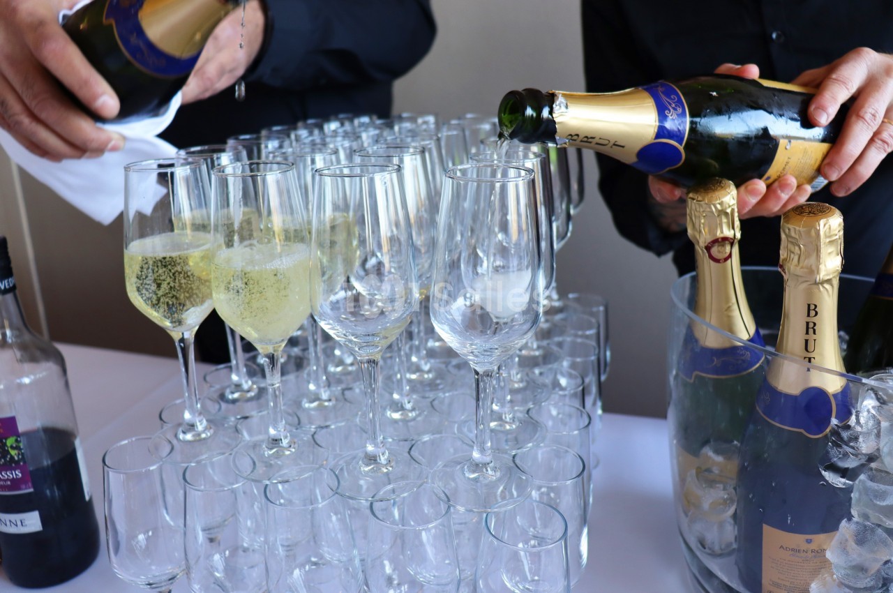 Deux personnes remplissent des flûtes avec du champagne, entourées de bouteilles et d'un seau à glace.