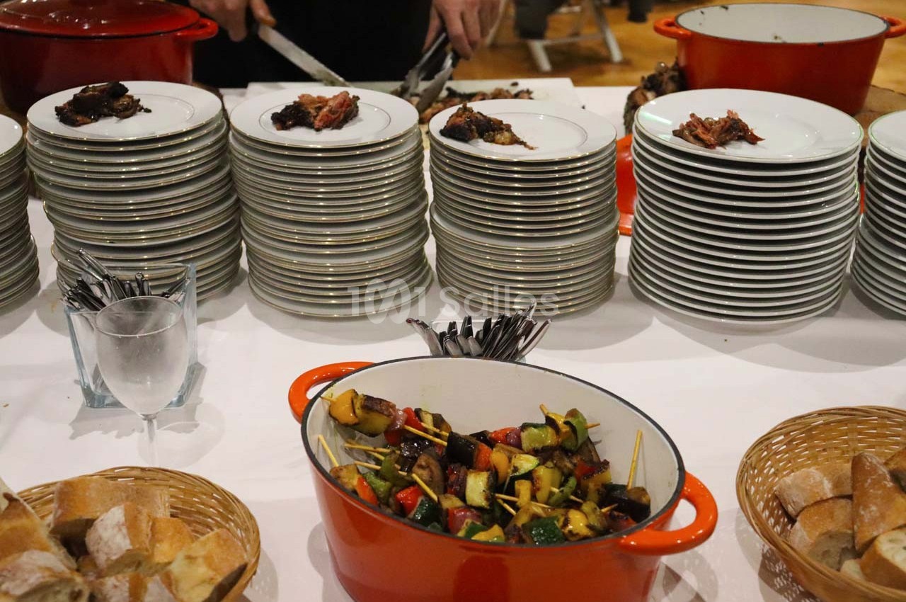 Assiettes empilées avec morceaux de viande, légumes grillés dans une cocotte, pain et couverts sur une table.