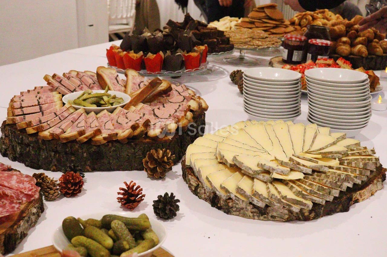 Plateau de charcuterie, fromage, cornichons et pains sur une table, entourés de desserts et vaisselle.