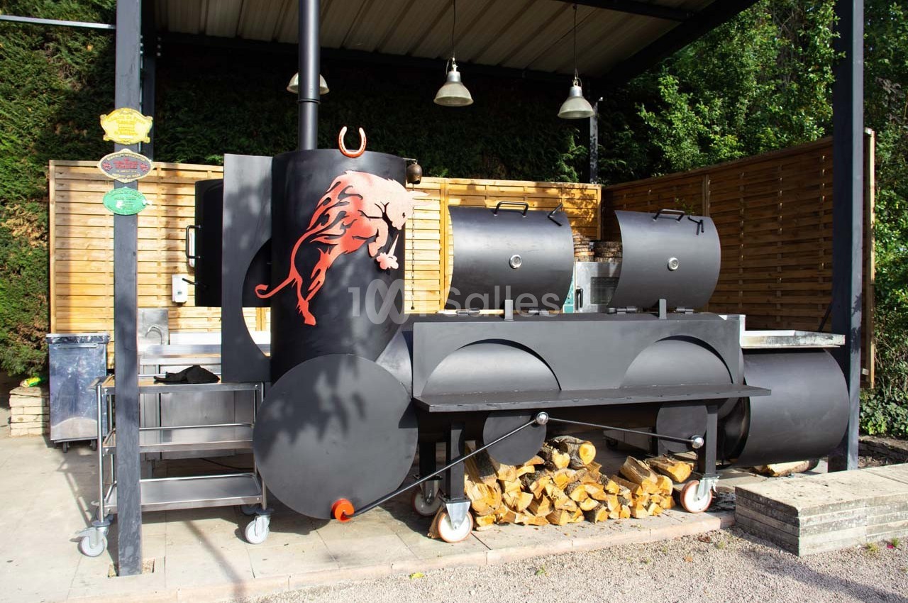 Barbecue fumoir noir en métal avec un motif de taureau rouge, abrité sous un auvent, entouré de bois de chauffage.