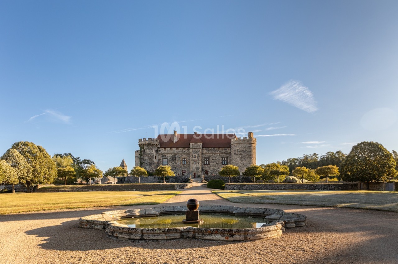 Vue d'un château en pierre avec un toit rouge, entouré d'arbres, et un bassin ornemental au premier plan.