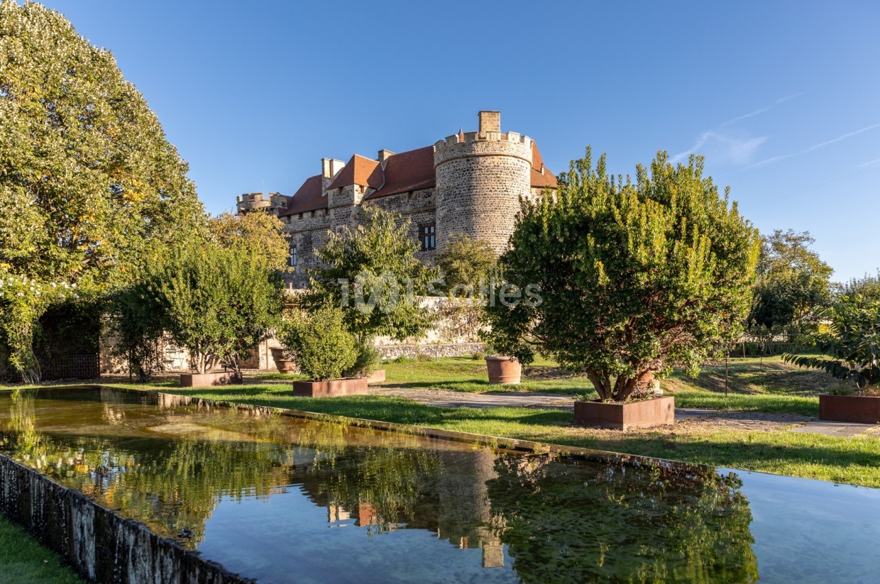 Vue d'un château en pierre entouré de jardins arborés, avec un bassin reflétant le bâtiment sous un ciel dégagé.