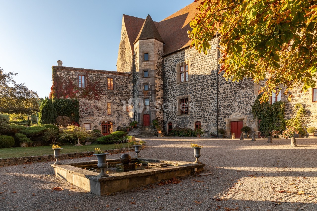 Façade en pierre d'un château historique avec un toit en tuiles, une fontaine et des arbres aux couleurs automnales.