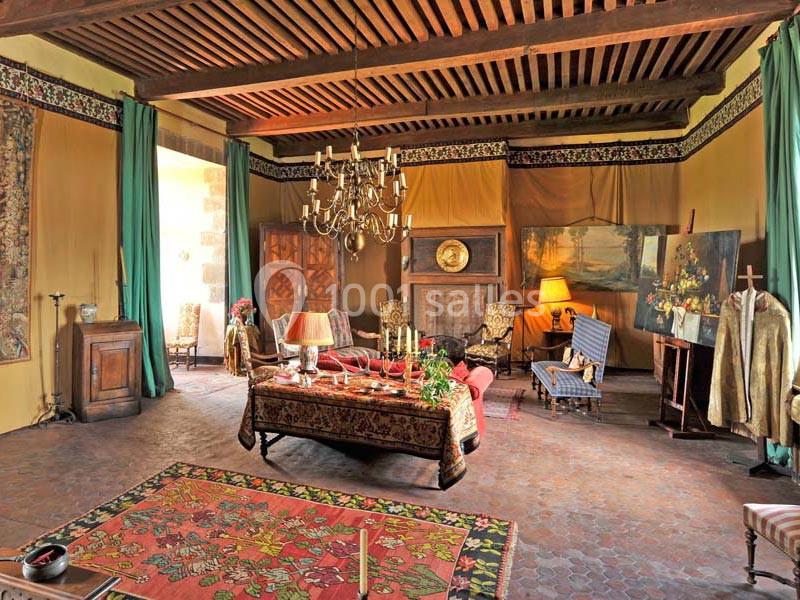 Salon ancien avec poutres apparentes, tapis coloré, mobilier classique, cheminée et éclairage chaleureux.