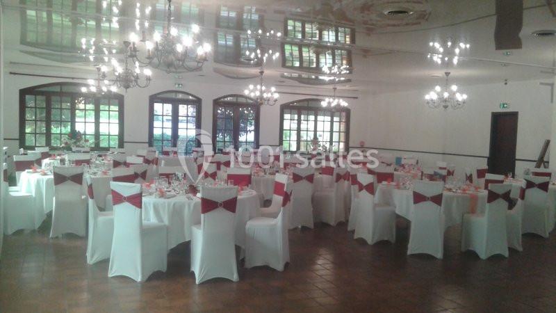 Salle de réception décorée avec des tables rondes dressées et des chaises blanches ornées de nœuds rouges.