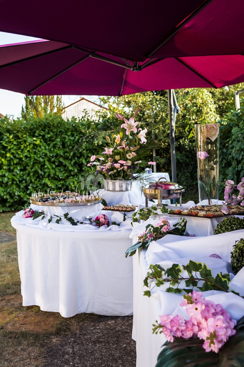 Buffet en plein air avec nappes blanches, fleurs roses et parasols violets, entouré de verdure.
