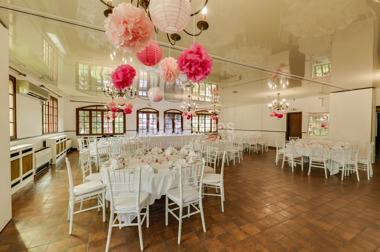 Salle de réception décorée avec des lanternes et pompons roses, tables rondes dressées avec nappes blanches.