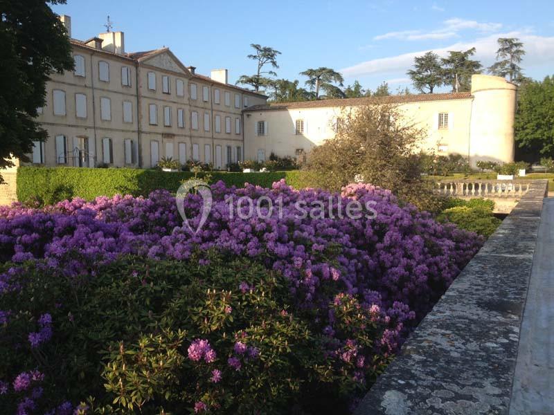 Massif de fleurs violettes devant un grand bâtiment en pierre à plusieurs étages, entouré de verdure.