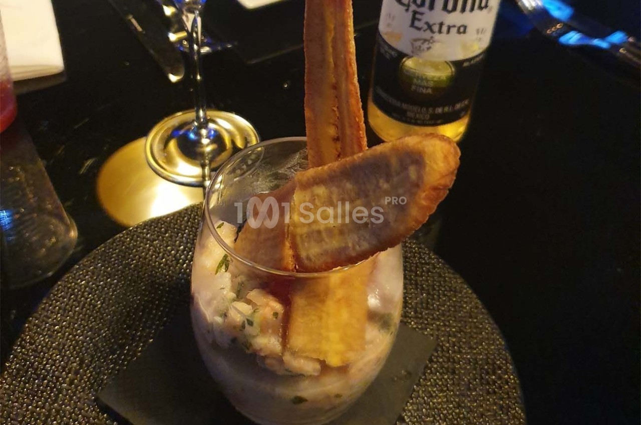 Verre de ceviche garni de chips de banane plantain, posé sur une serviette noire avec des verres en arrière-plan.