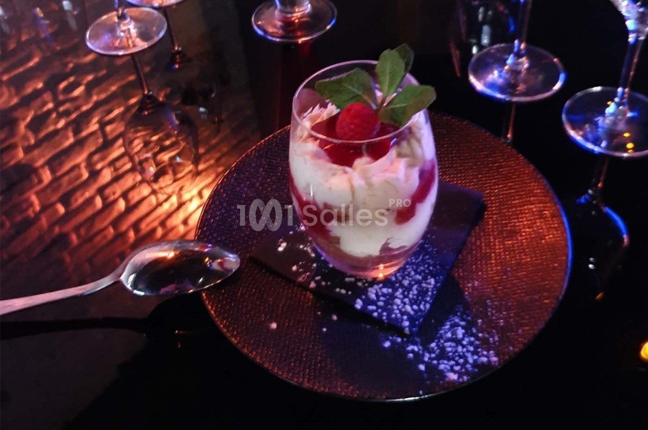 Verre de dessert composé de crème, framboises et feuilles de menthe, posé sur une assiette noire avec une cuillère.