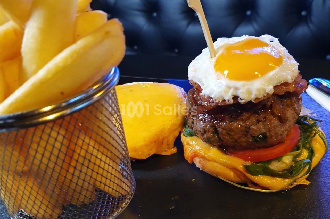 Burger garni d'un œuf au plat, bacon, légumes et fromage, accompagné de frites dans un panier métallique.
