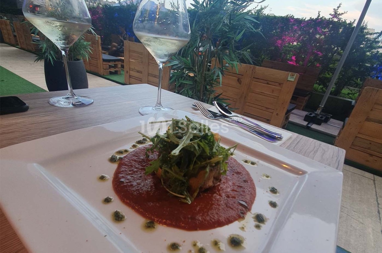 Assiette de plat dressé avec une sauce rouge, garnie de roquette, servie en terrasse avec deux verres de vin blanc.