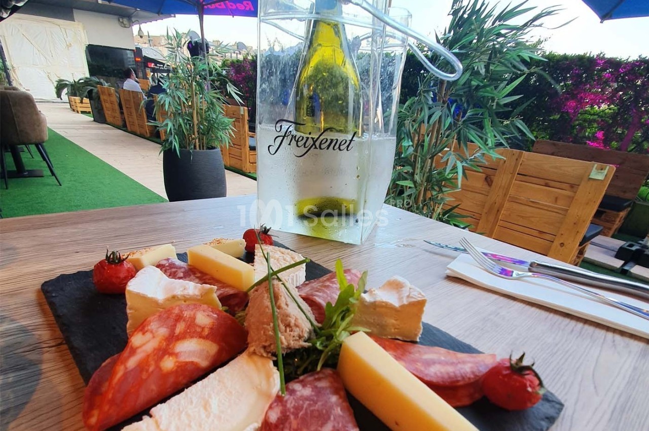 Planche de charcuterie et fromages sur une table en terrasse, avec une bouteille de vin blanc dans un seau à glace.