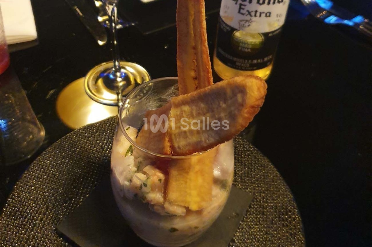 Un verre contenant un ceviche garni de chips de banane plantain, posé sur une table sombre avec des verres en arrière-plan.