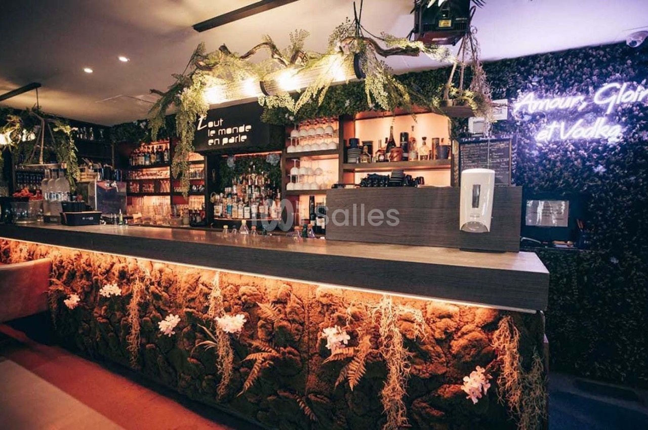 Bar moderne avec comptoir éclairé, décor végétal, étagères de bouteilles et néon affichant ’Amour, Gloire, Vodka’.